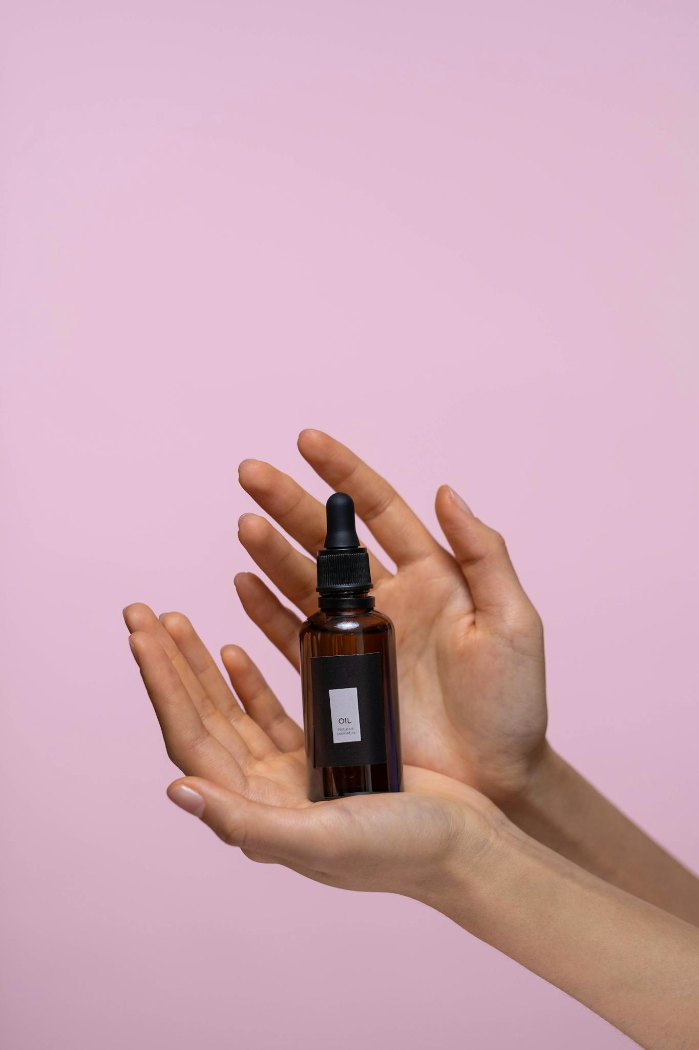 Close-up of a brown dropper bottle held by hands against a pink background, representing natural skincare.