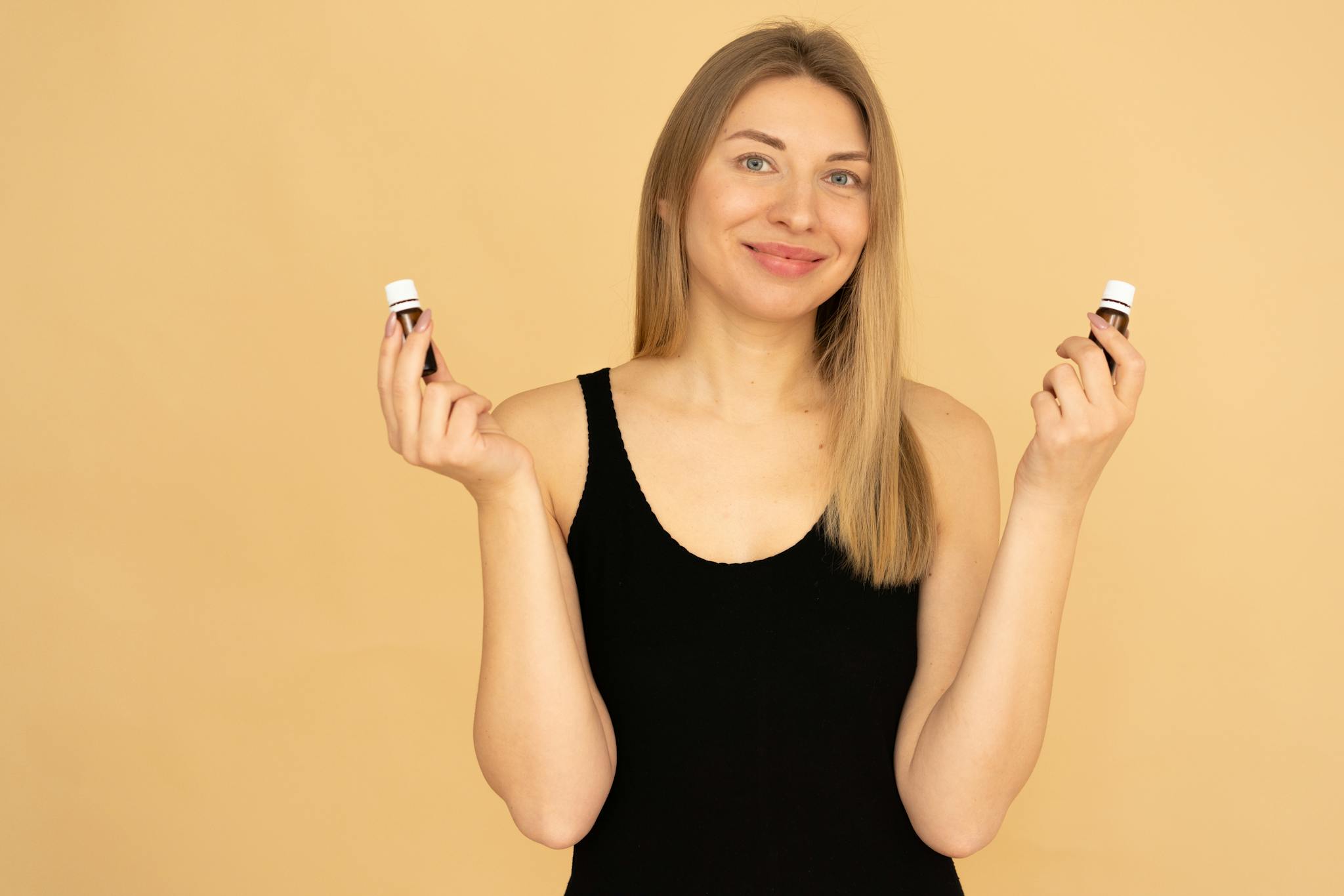 Woman holding essential oils with a joyful expression against a beige background, emphasizing wellness and beauty.